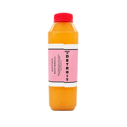 A bottle of apple ginger tea, featuring a bright pink label and a red cap, filled with orange-colored liquid, set against a plain white background.